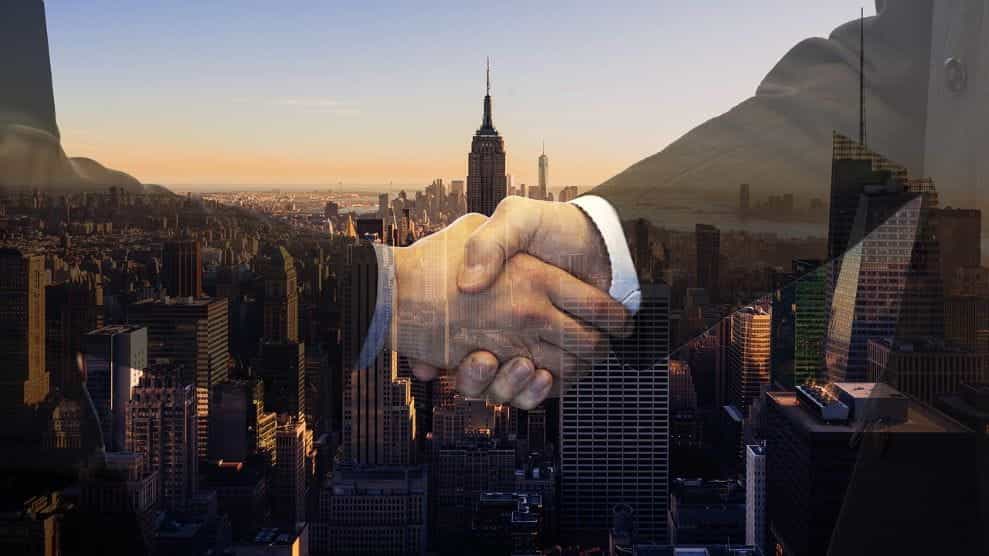 Two translucent figures shaking hands in front of a backdrop showing the skyline of midtown Manhattan in New York City.