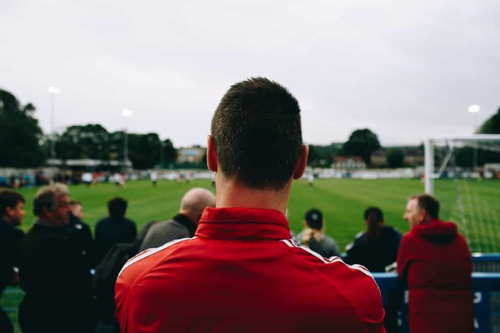 Seorang pria berjaket merah menonton pertandingan sepak bola dari pinggir lapangan.