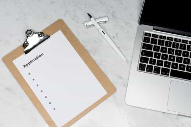 Kertas putih di papan klip dengan kata aplikasi sebagai judulnya, diletakkan di sebelah laptop.