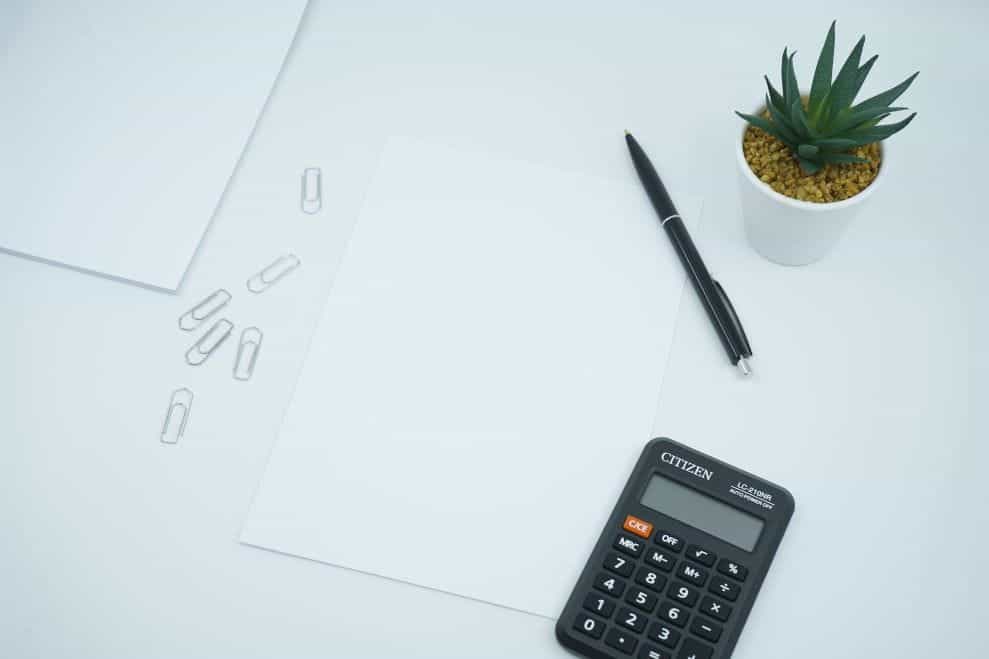 A black calculator placed next to white paper, some clips, and a pot.