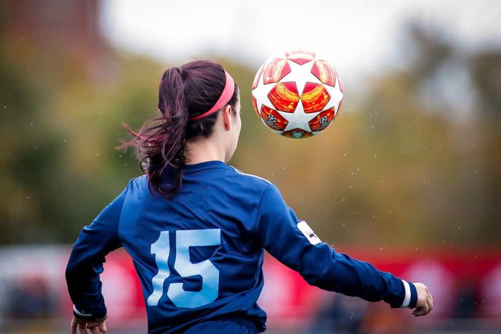 Dari belakang, seorang wanita menendang bola sepak ke udara.