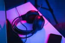 A gamer’s headphones placed next to a phone on a desk, under pink lights.