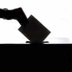 A silhouetted hand posts a vote into a ballot box.