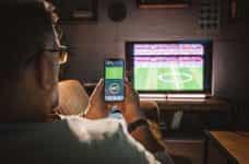 A man sits on a sofa watching a soccer game and using a smart phone to place a bet.