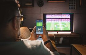 A man sits on a sofa watching a soccer game and using a smart phone to place a bet.