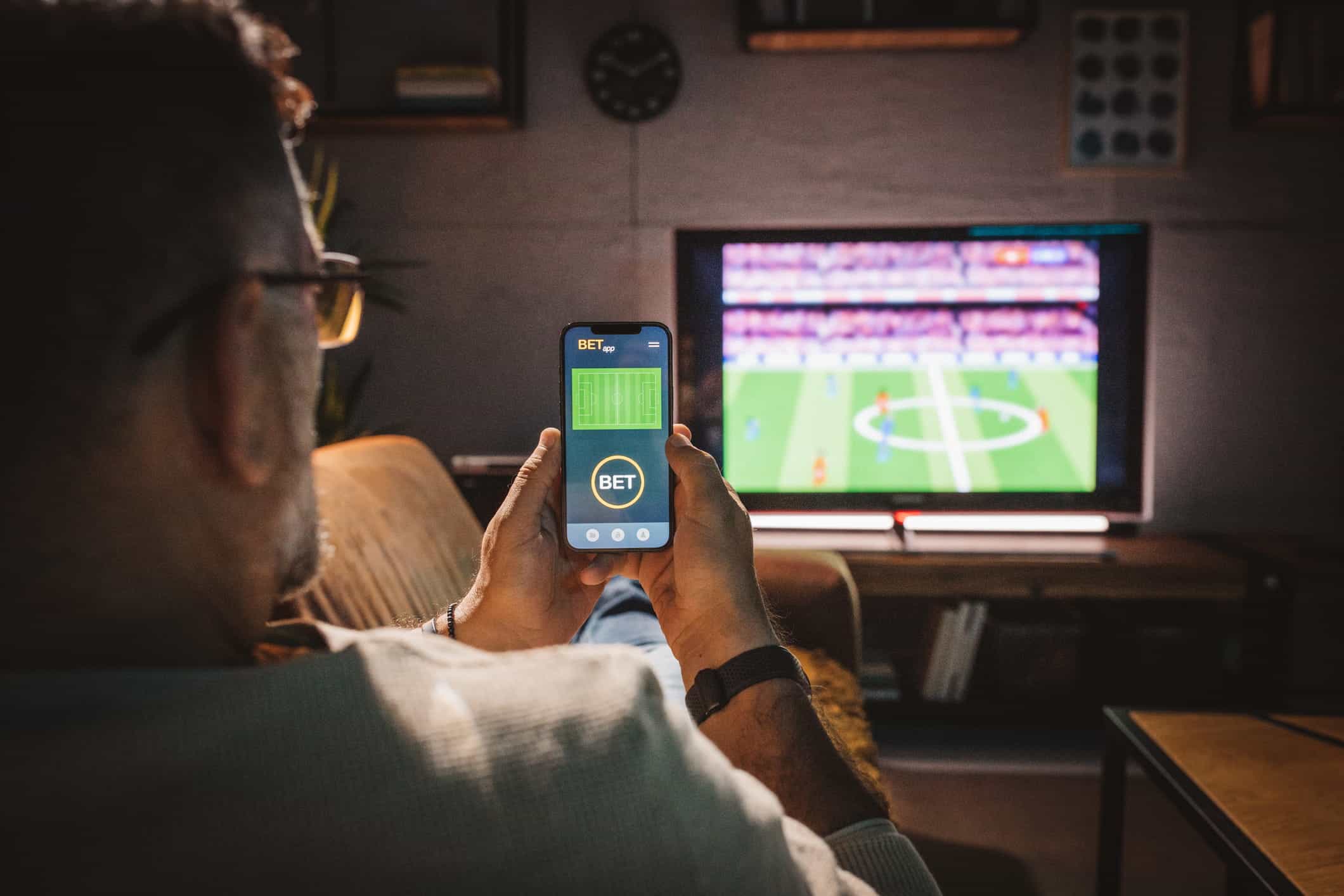Seorang pria duduk di sofa menonton pertandingan sepak bola dan menggunakan ponsel pintar untuk memasang taruhan.