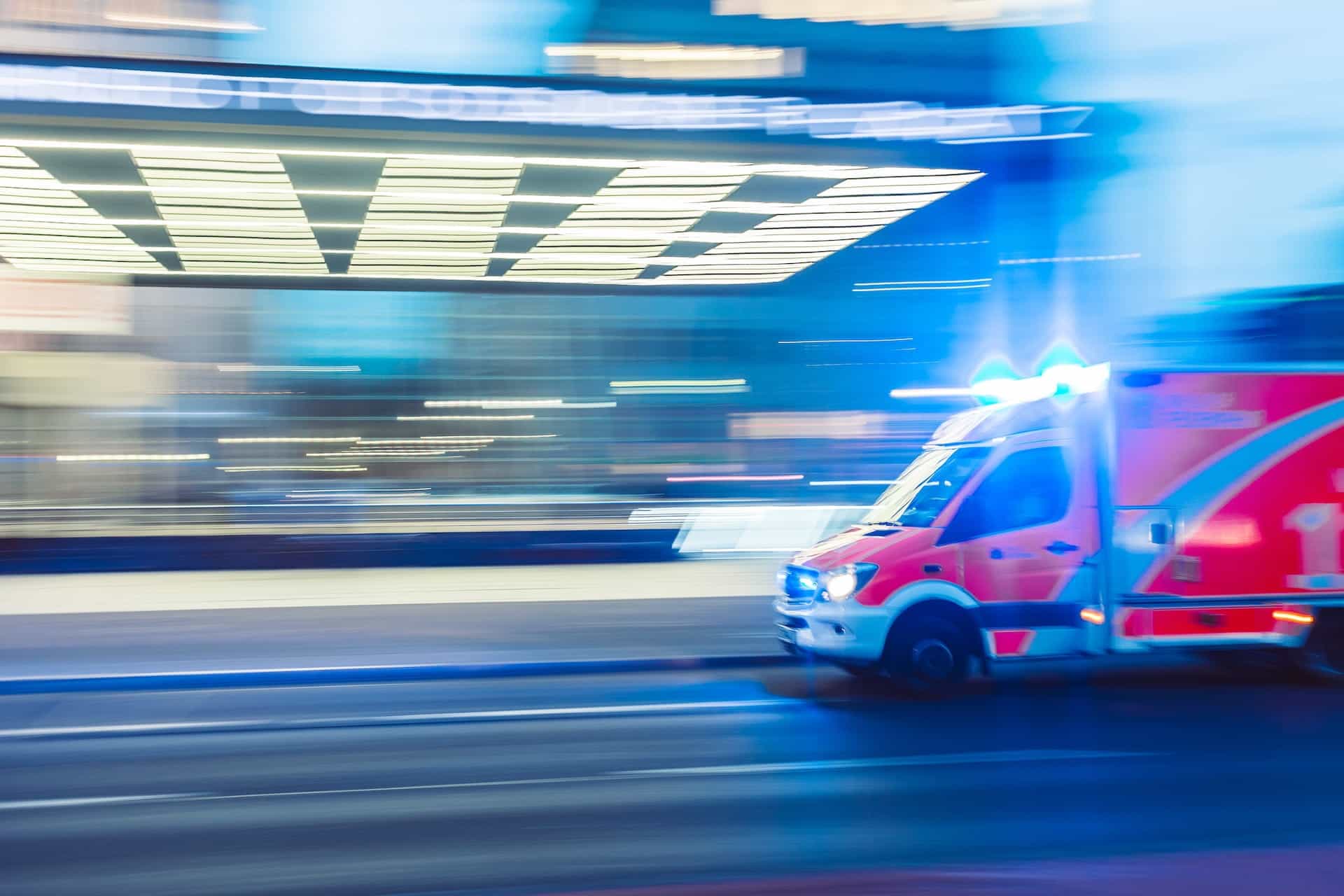 An ambulance with its light flashing drives in front of a brightly lit building.