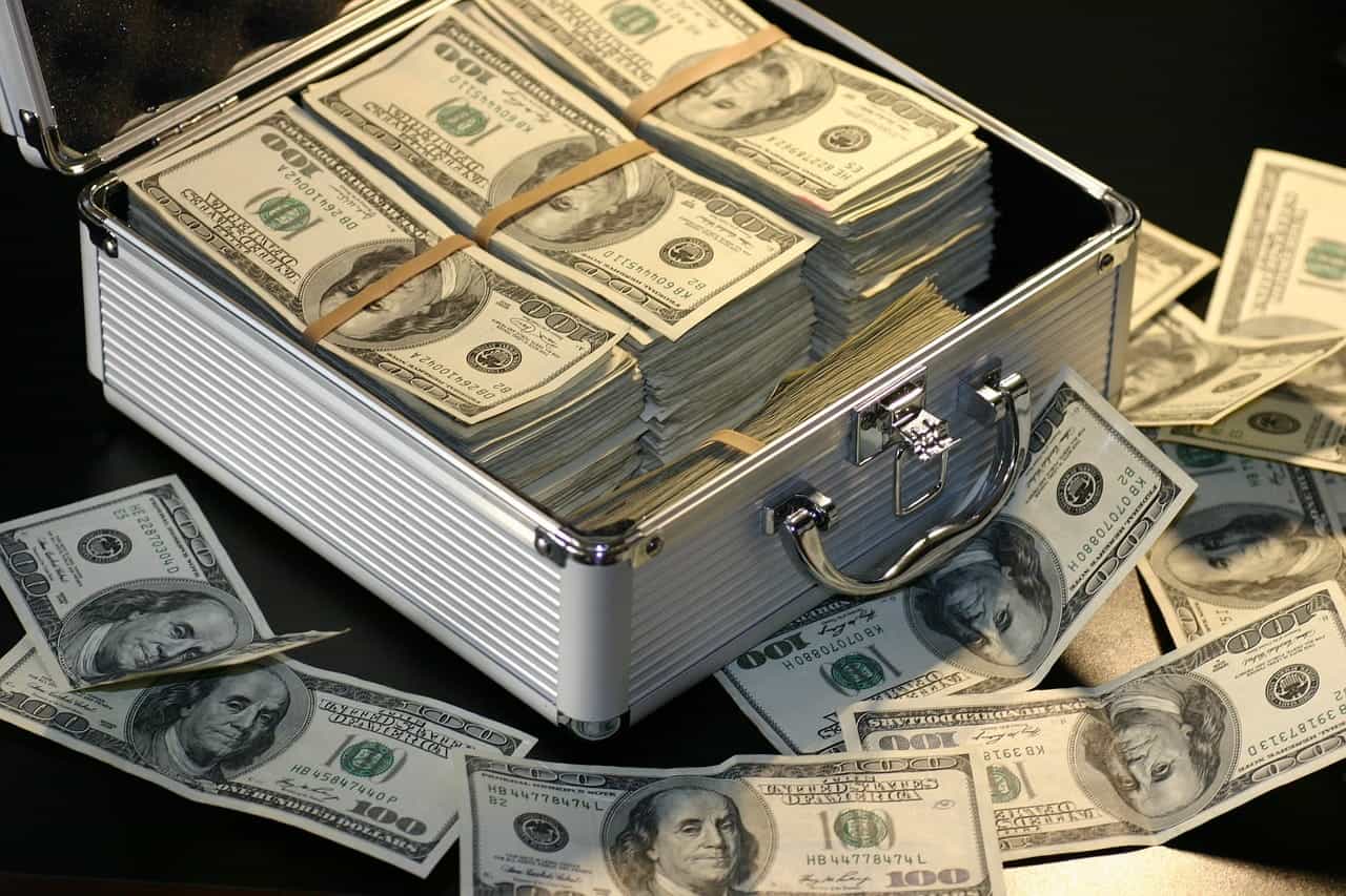 A thick silver briefcase with stacks of $100 US Dollar bills sitting in it, as well as several other single $100 US Dollar bills scattered around it.