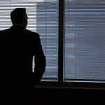 The dark silhouette of a man in a suit looking through the thin blinds of the windows in his office.