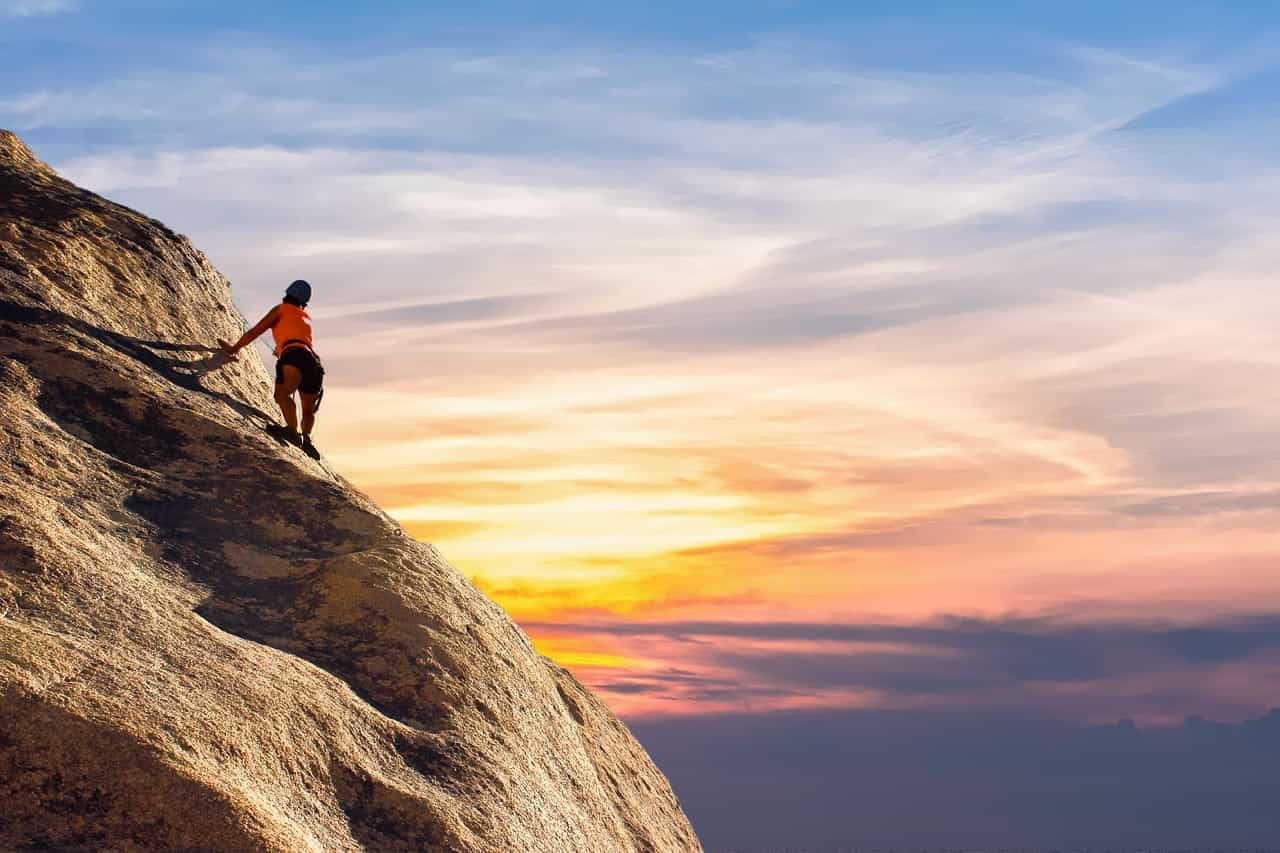 A woman climbing the side of a rocky mountain, with cloud-whipped sky featuring burning red sun streaks stretching out in front of her.