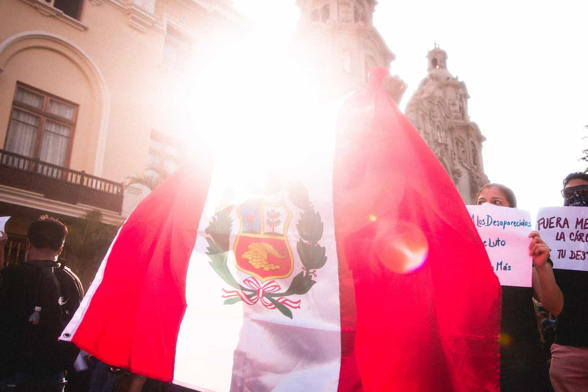 Bendera Peru melambai di depan kerumunan yang berbaris, diterangi oleh sinar matahari di belakangnya. 