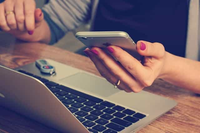 Seorang wanita memegang smartphone sambil duduk di depan laptop terbuka yang memiliki stik USB yang diletakkan di permukaannya tepat di sebelah keyboardnya.