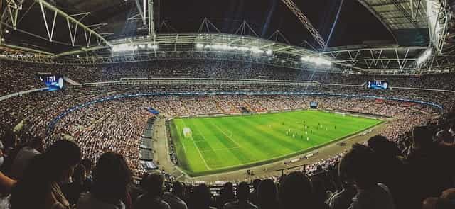 Stadion sepak bola yang penuh sesak selama pertandingan sepak bola, dengan lapangan hijau di tengahnya, diterangi oleh lampu sorot yang terang.