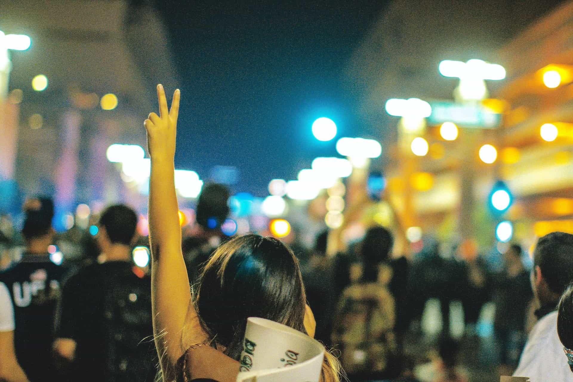 Dari belakang, seorang wanita mengangkat tangannya dengan tanda perdamaian dalam protes jalanan di malam hari.