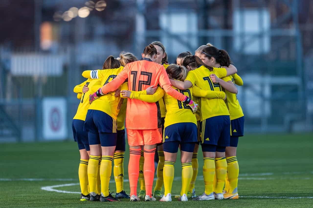Sebuah tim pemain sepak bola wanita berkerumun dalam lingkaran untuk berkonsultasi satu sama lain mengenai strategi dan taktik sebelum pertandingan dimulai.