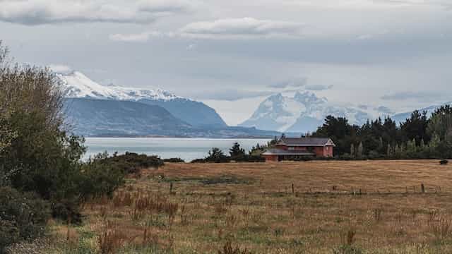 Sebuah rumah di lapangan di pantai Puerto Natales, Chile, dengan pegunungan bersalju di kejauhan.