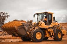 A yellow bulldozer pauses in the midst of reddish sand, driven by someone in an orange uniform.