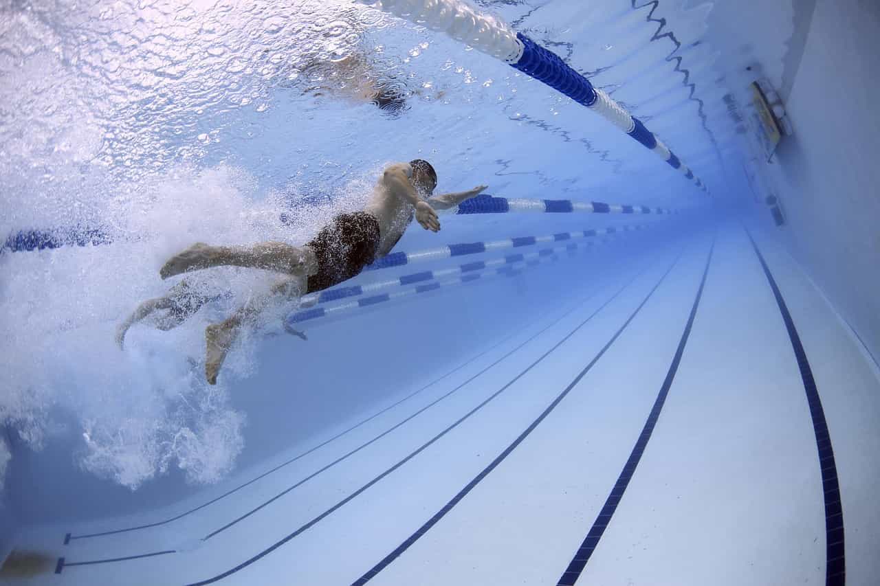 Seorang perenang melakukan gerakan merangkak depan di bawah air sambil berenang di jalurnya di kolam renang ukuran Olimpiade, dengan perenang lain di jalur sebelah kirinya.