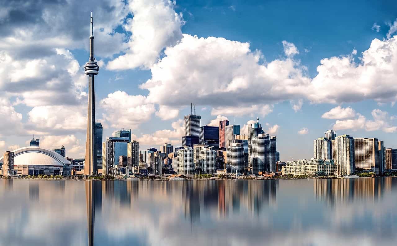 Cakrawala pusat kota Toronto, Ontario pada siang hari, menampilkan beberapa gedung tinggi dan gedung pencakar langit, serta Menara CN yang terkenal di dunia, semuanya tercermin di permukaan Danau Ontario di latar depan.