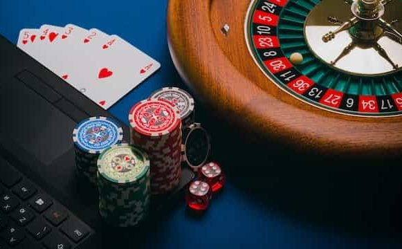 A roulette wheel, playing cards, poker chips and dice with a laptop.