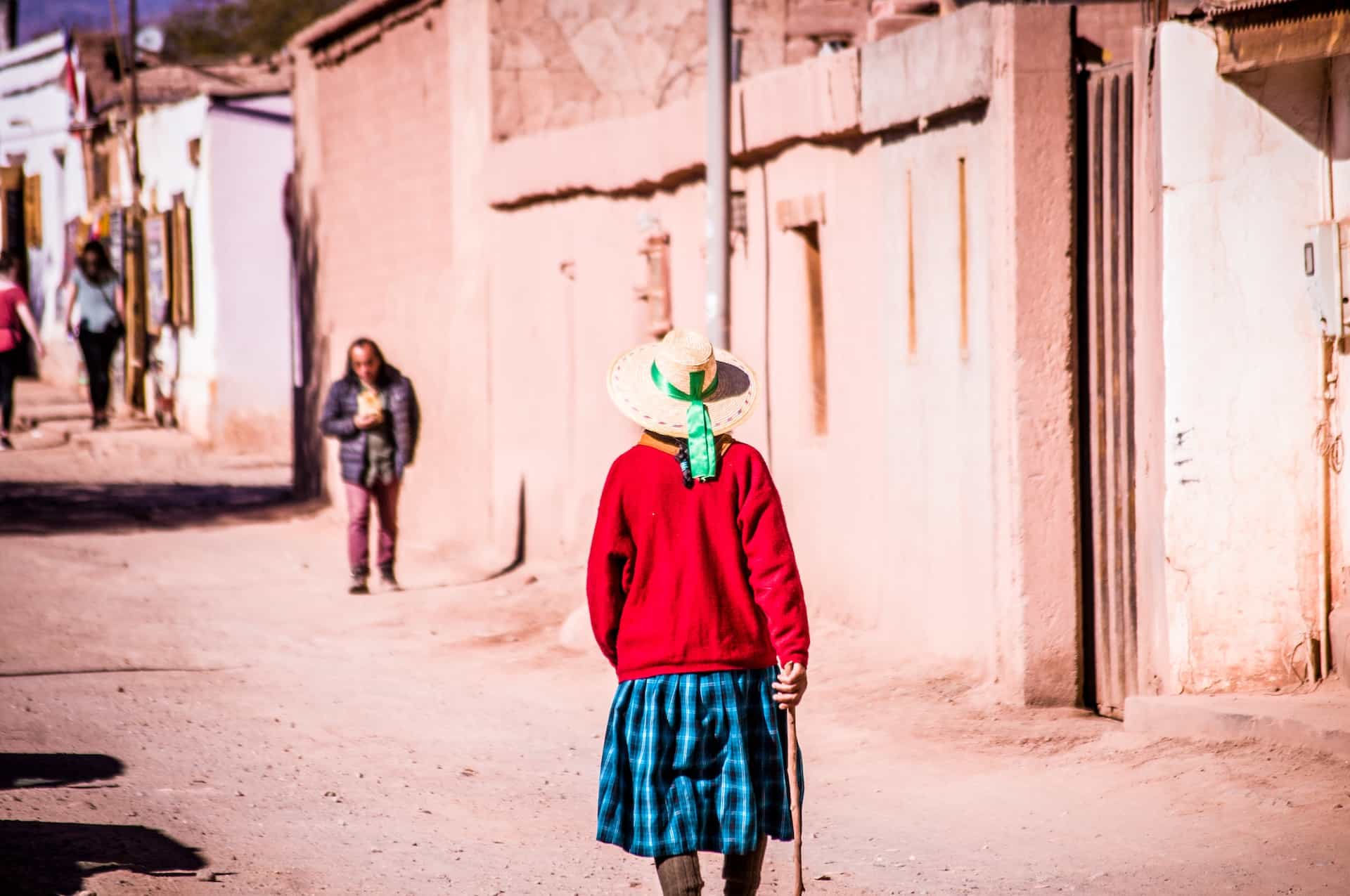 A woman walking with a cane, a hat and a red sweater in San Pedro de Atacama, Chile.