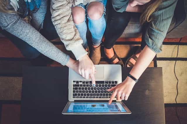 Tiga orang duduk di depan laptop yang diletakkan di atas meja, dengan dua orang menunjuk ke sesuatu di layar laptop sementara orang ketiga mengarahkan kursor menggunakan touch pad laptop.