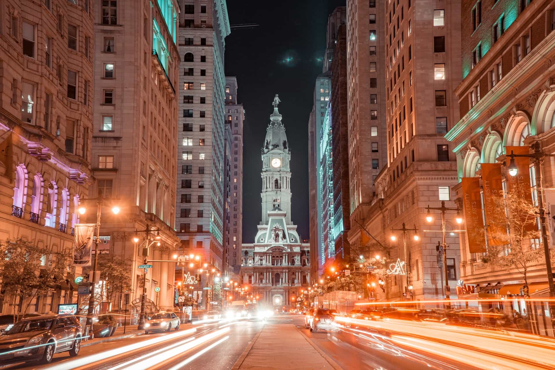 Sebuah jalan yang sibuk di pusat kota Philadelphia, Pennsylvania, menampilkan bidikan lalu lintas yang berlalu-lalang dengan eksposur panjang dan menara jam yang tinggi di ujung jalan.