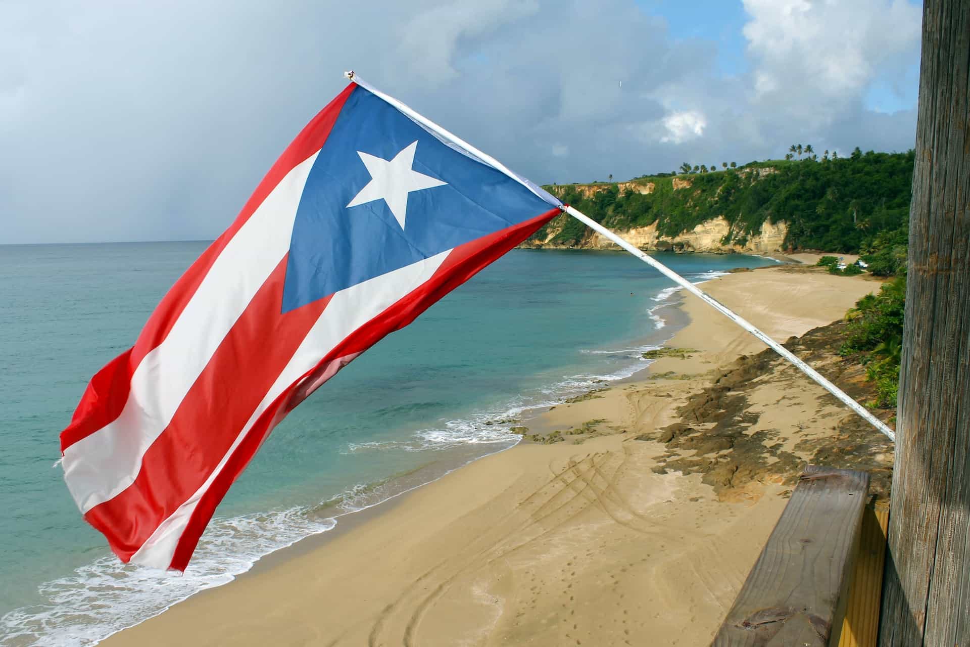 Bendera Puerto Rico berkibar di atas pantai berpasir, dengan tebing yang tertutup pepohonan dan samudra biru terbentang di belakangnya.