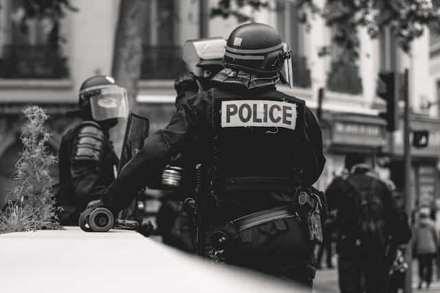 Gambar hitam putih polisi dengan alat pelindung menunggu di sebuah acara di Lyon, Prancis.