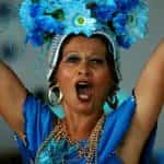 A woman in a sleeveless blue dress and elaborate, flower headdress dances and shouts at Carnival in Sao Paulo, Brazil.
