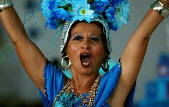 A woman in a sleeveless blue dress and elaborate, flower headdress dances and shouts at Carnival in Sao Paulo, Brazil.