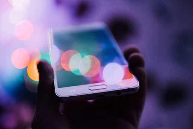 A person’s hand holding up a smartphone in horizontal orientation with lights refracting off of its screen.