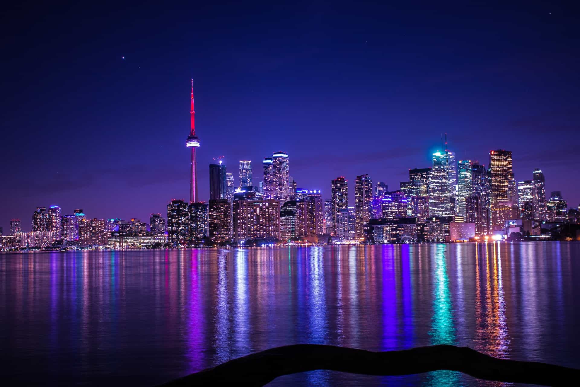 Cakrawala pusat kota Toronto, Ontario yang ramai di Kanada, menampilkan lusinan gedung pencakar langit yang terang dan gedung-gedung tinggi serta Menara CN yang ikonis, dengan Danau Ontario terbentang di depannya.