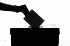 A silhouette of a person’s hand slipping an envelope into a ballot box during an election.