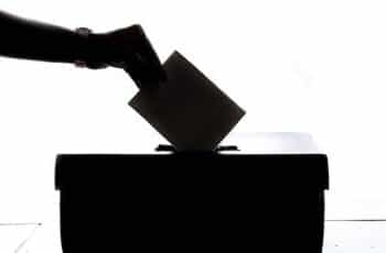 A silhouette of a person’s hand slipping an envelope into a ballot box during an election.