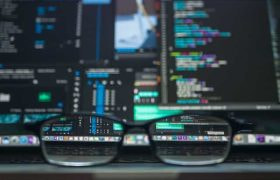 A pair of glasses placed in on the surface of a laptop, the screen of which is displaying several lines of code and text.