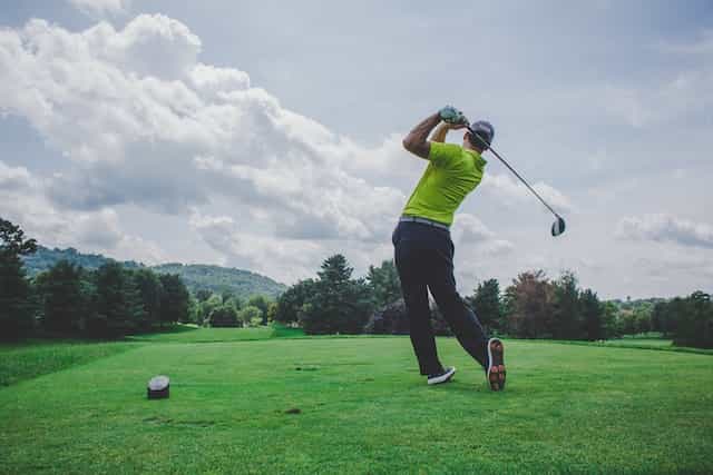 A male golfer post-golf swing on an outdoor golf course, with rolling green mountains seen off in the distance.