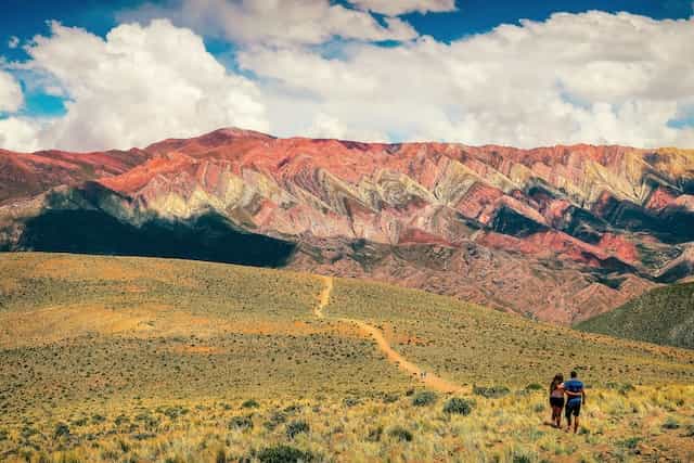 A couple walks up a hill covered in green and yellow shrubs at the base of red mountains in Jujuy, Argentina.