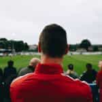 The back of a man’s head while he watches a soccer game from the stands at an outdoor soccer field.