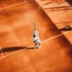 A man in a white tennis outfit serves a ball standing on an orange clay court.