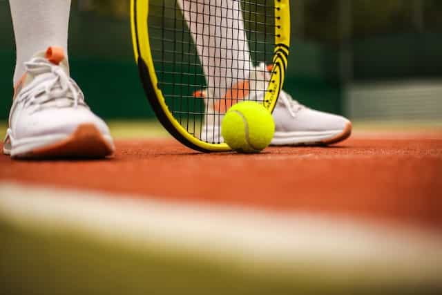 A tennis player’s racket resting on the ground of a clay outdoor tennis court with a tennis ball sitting in front of it.