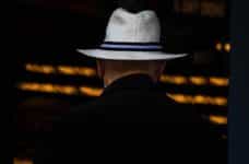 The back of a person dressed in a black suit and in a white hat with a blue strip running around it.
