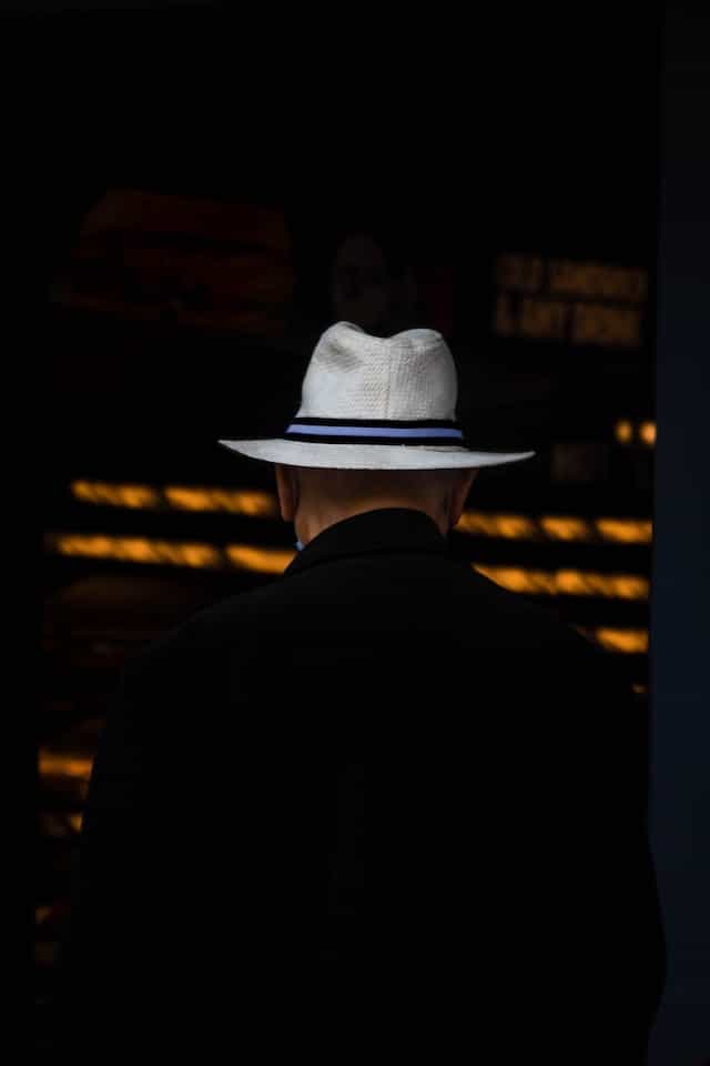 The back of a person dressed in a black suit and in a white hat with a blue strip running around it.