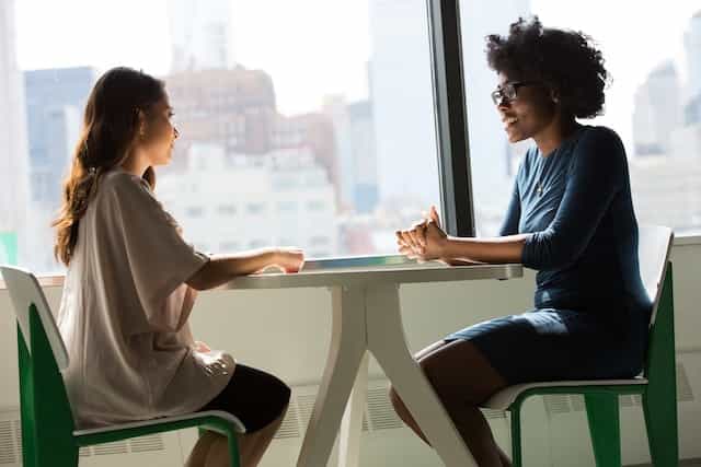 Two women sitting at a small table in an office environment while speaking to one another.