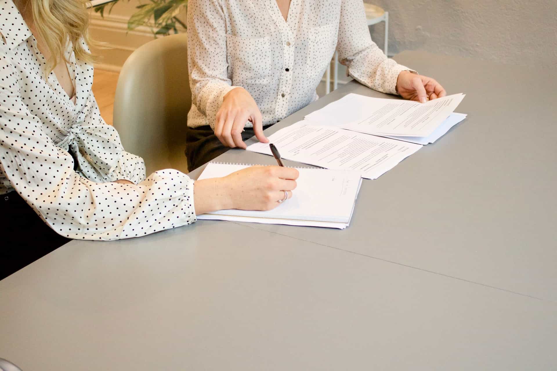 A person scribbling on white paper beside a person with documents.
