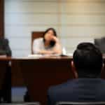 From behind, a man sits in front of a panel of three observers in a courtroom.