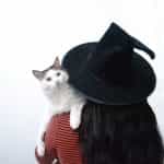 A woman with long hair wears a witches hat and holds a cat on her shoulder.