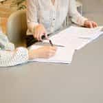 Two women sitting at a desk in an office reviewing paperwork and signing a written agreement.