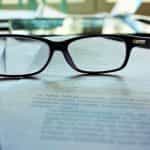 A pair of glasses resting atop a pile of paperwork placed on a table.