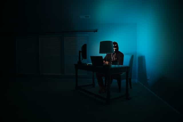 A masked and shadowy figure sitting at a desk in front of two computer monitors while being drenched in blue light.